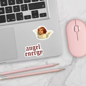 May include: A silver laptop computer with a pink mouse and two pink pencils on a white marble surface. The laptop has a sticker of an angel with the text "angel energy" in red gothic font.