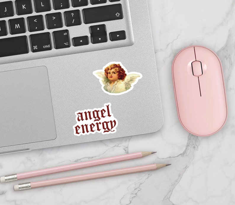 May include: A silver laptop computer with a pink mouse and two pink pencils on a white marble surface. The laptop has a sticker of an angel with the text "angel energy" in red gothic font.