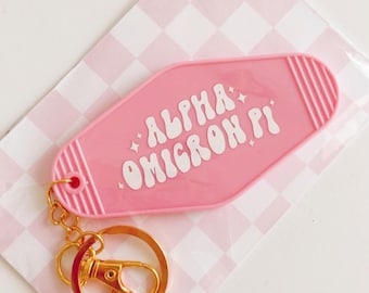 Może przedstawiać: Różowy brelok do kluczy w kształcie klucza hotelowego z złotym kółkiem. Brelok ma napis "ALPHA OMICRON PI" w kolorze białym.