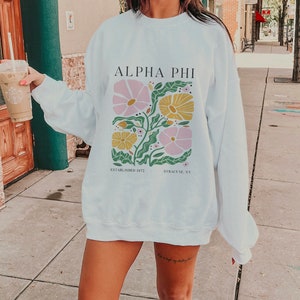 May include: White sweatshirt with a floral design and the text "Alpha Phi Established 1972 Syracuse, NY".