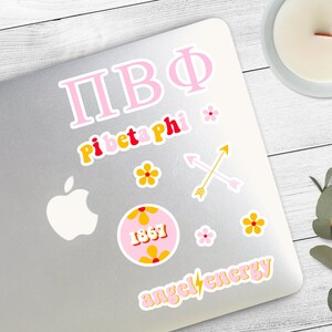 May include: A silver laptop with a variety of stickers. The stickers are pink, yellow, and red. The stickers include the letters "ПВФ", "Pi Beta Phi", a flower, a circle with the number "1867", two crossed arrows, and the words "angel energy".
