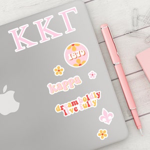 Pode incluir: Um laptop prateado com adesivos rosa e amarelos. Os adesivos incluem as letras "KKG", um círculo com o ano "1870" dentro, a palavra "kappa" e a frase "dream boldly live fully".