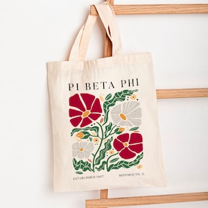 Könnte beinhalten: Eine weiße Canvas-Tragetasche mit einem floralen Design in Rot, Weiß und Grün. Die Tasche trägt den Text "Pi Beta Phi" in Schwarz, "Established 1867" in Schwarz und "Monmouth, IL" in Schwarz.
