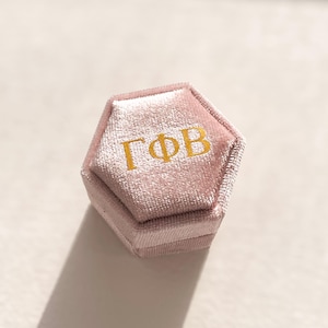 Könnte beinhalten: Eine sechseckige, rosa Samt-Ringbox mit goldfarbenen Buchstaben. Die Box ist geschlossen, mit Deckel und Boden. Die goldenen Buchstaben auf dem Deckel lauten "ΓΦB". Die Box steht auf einer weißen Oberfläche.