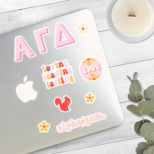 May include: A laptop sticker set featuring the letters "AGD" in pink, a pink circle with the text "1994" and yellow flowers, a red squirrel, and the text "alpha gam" in yellow with a lightning bolt.