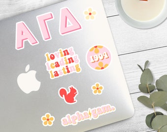 May include: A laptop sticker set featuring the letters "AGD" in pink, a pink circle with the text "1994" and yellow flowers, a red squirrel, and the text "alpha gam" in yellow with a lightning bolt.