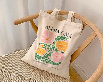 May include: A beige canvas tote bag with a floral design in pink, yellow, and green. The bag has the text "Alpha Gam" in black lettering and "Established 1904 Syracuse, NY" in smaller black lettering.