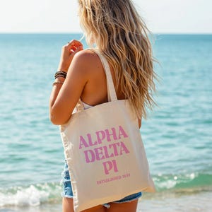 May include: A beige canvas tote bag with the words "ALPHA DELTA PI" in pink block letters. Below the text is "ESTABLISHED 1851" in a smaller pink font. The bag is carried by a person on a beach.