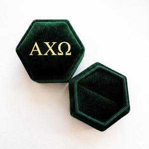 Op de afbeelding: Zeshoekige donkergroene fluwelen ringdoos met gouden letters "AX&Omega;". De doos is open en onthult de binnenkant. De doos staat op een wit oppervlak.