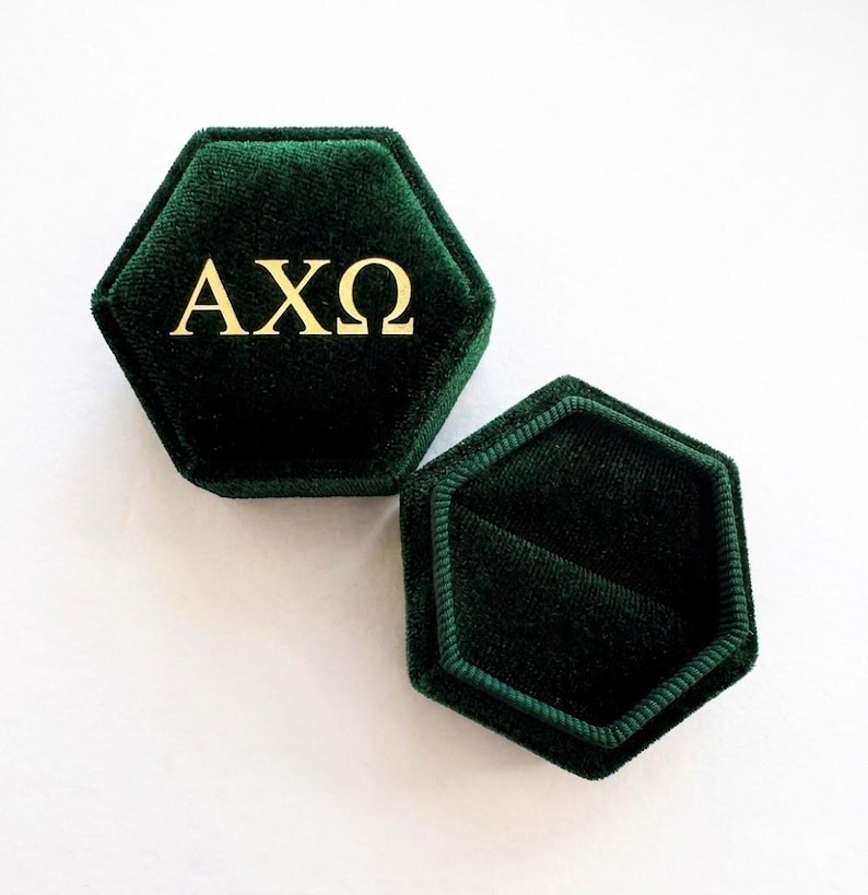 Op de afbeelding: Zeshoekige donkergroene fluwelen ringdoos met gouden letters "AX&Omega;". De doos is open en onthult de binnenkant. De doos staat op een wit oppervlak.