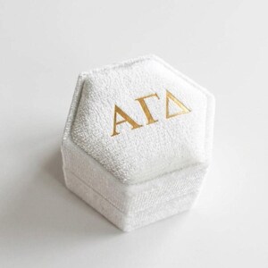 May include: A white, hexagonal ring box with a textured surface. The box has gold lettering that reads "&Alpha;&Gamma;&Delta;" on the top. The box is closed and sits on a white background.
