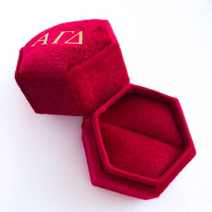 May include: Open red velvet ring box with gold lettering "&Alpha;&Gamma;&Delta;" on the lid. The box is hexagonal and has a soft, plush interior. The box is open, revealing the inside, and sits on a white surface.