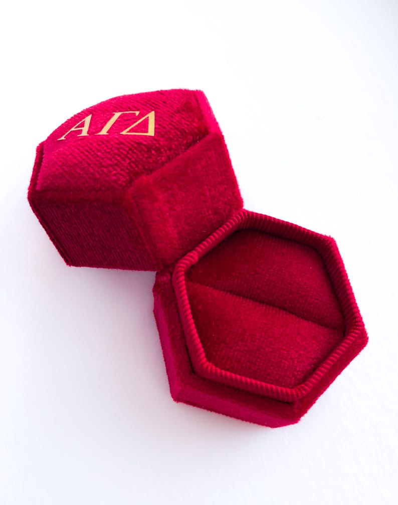 May include: Open red velvet ring box with gold lettering "&Alpha;&Gamma;&Delta;" on the lid. The box is hexagonal and has a soft, plush interior. The box is open, revealing the inside, and sits on a white surface.