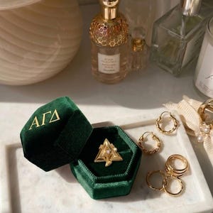 May include: A gold ring with a triangle design and a small gemstone is displayed in an open, dark green velvet ring box. Several gold rings and hoop earrings are arranged on a white marble tray, alongside perfume bottles.
