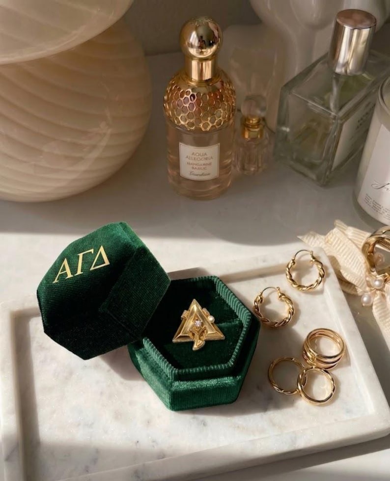 May include: A gold ring with a triangle design and a small gemstone is displayed in an open, dark green velvet ring box. Several gold rings and hoop earrings are arranged on a white marble tray, alongside perfume bottles.