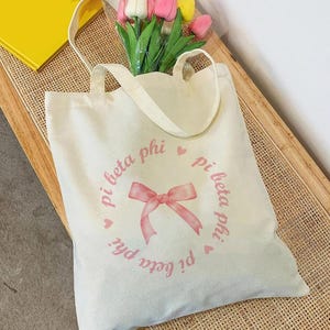 Könnte beinhalten: Eine weiße Canvas-Tragetasche mit rosa Schriftzug "pi beta phi" und einer rosa Schleife. Die Tasche hat zwei Henkel.