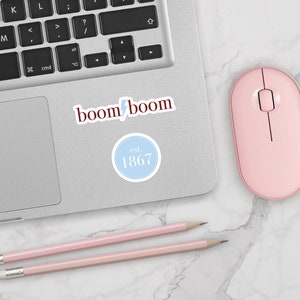 May include: A laptop computer with a pink mouse and two pink pencils on a white marble surface. The laptop has a sticker that says "boom boom" and another sticker that says "est. 1867".