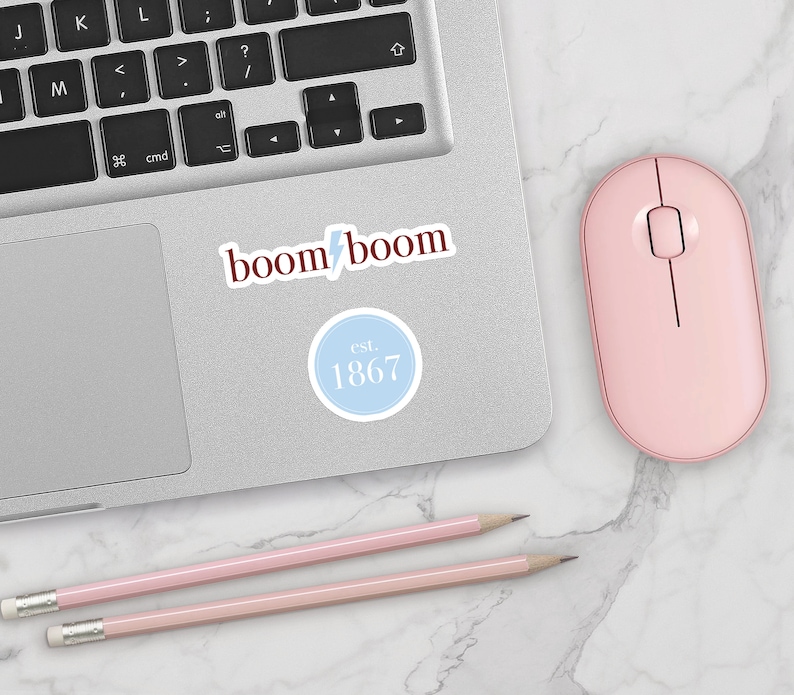 May include: A laptop computer with a pink mouse and two pink pencils on a white marble surface. The laptop has a sticker that says "boom boom" and another sticker that says "est. 1867".