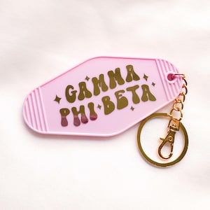 May include: A pink motel-style keychain with gold lettering spelling "GAMMA PHI BETA". The keychain has a gold-coloured key ring and clasp. The background is a soft, white fabric.