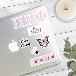 May include: A laptop computer with several pink and black stickers. The stickers include the letters "ПВФ", "est. 1867", "ring ching", a butterfly with the text "pi beta phi", two stars, and the text "pi beta phi".