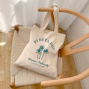 Könnte beinhalten: Eine beige Canvas-Tragetasche mit einem grünen Palmenbaum-Design und dem Text "Pi Beta Phi Women's Fraternity Established 1867".