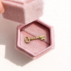 May include: A small gold-toned key charm featuring the Greek letters KKT and AΩ engraved on its shaft. The key is resting inside a hexagonal, dusty pink velvet jewelry box with the lid partially open, revealing the soft interior lining.