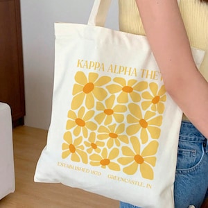 May include: A white canvas tote bag with a yellow daisy pattern and the text "Kappa Alpha Theta Established 1870 Greencastle, IN".