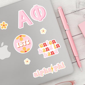 May include: A laptop computer with several pink and yellow stickers. The stickers feature the letters "AΦ", the year "1872", the phrase "union hand in hand", and the words "alpha phi" with a lightning bolt.