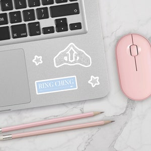 May include: A laptop computer with a pink mouse and two pink pencils on a white marble surface. The laptop has a sticker with the text "RING CHING" and a silver glitter star sticker.