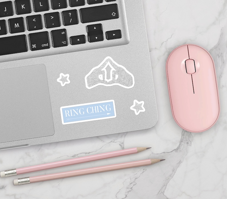 May include: A laptop computer with a pink mouse and two pink pencils on a white marble surface. The laptop has a sticker with the text "RING CHING" and a silver glitter star sticker.