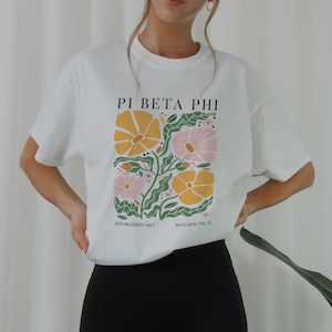 Könnte beinhalten: Weißes T-Shirt mit einem Blumendesign und dem Text "Pi Beta Phi Established 1867 Monmouth, IL"