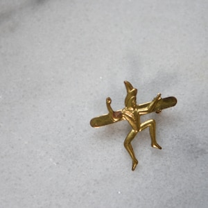 Pode incluir: Um alfinete de metal dourado com uma figura estilizada de uma pessoa com um chapéu pontudo, segurando um pau e em pé sobre uma barra horizontal.