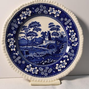 May include: A blue and white ceramic plate with a scalloped edge. The plate features a blue and white floral pattern and a scene of a bridge over a stream with trees and a house in the background.