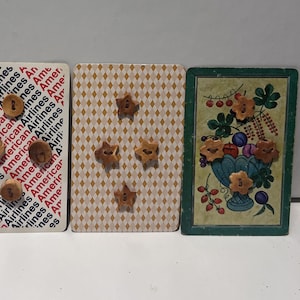 Puede incluir: Cuatro naipes con botones de madera adjuntos. Las cartas presentan varios diseños, incluyendo un patrón de caras sonrientes amarillas, texto de American Airlines, un patrón de diamantes y una ilustración floral.