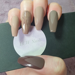May include: A set of five long, almond-shaped false nails with a brown, beige and marble design. The nails are displayed on a black box with a white circle that says "LCW Nails".