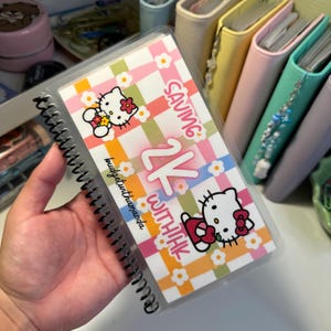 May include: A spiral-bound savings tracker featuring a colourful checkered pattern in pink, orange, and green with white flowers. The text reads "SAVING 2K WITH HK" and "budgetwithamanda" alongside two illustrations of a white cat character.
