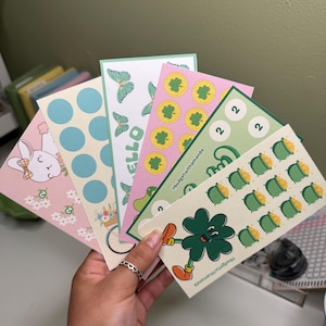 May include: A hand holds a collection of colorful sticker sheets. Designs include a bunny, polka dots, butterflies, shamrocks, and pots of gold. The text "HELLO" and "#budgetwithamanda" are visible on some sheets.