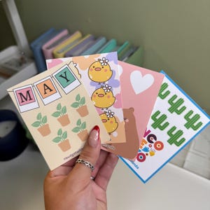 May include: Four colorful stickers with different designs. The first sticker is yellow with a repeating pattern of a chick with a flower crown. The second sticker is light pink with a white heart. The third sticker is yellow with a banner that says "MAY" in pink, green, and yellow letters. The fourth sticker is blue with a repeating pattern of green cacti and the text "HELLO" in colorful letters.