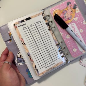 Op de afbeelding: Een lila planner met een budget tracking inzet. De inzet heeft kolommen voor categorie, datum, +/- en saldo. Een zwarte stift en een roze kaart met een tekenfilmprinses zijn ook zichtbaar. De planner heeft stickers en de tekst "BUDGETWITHAMANDA".