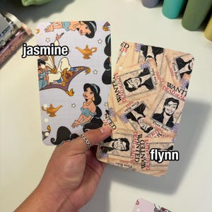 Op de afbeelding: Twee decoratieve plannerinzetstukken worden in een hand gehouden. E&eacute;n toont het personage Jasmine uit Aladdin, met de tekst "jasmine". De andere heeft een "Wanted Dead or Alive"-posterontwerp met de tekst "flynn".