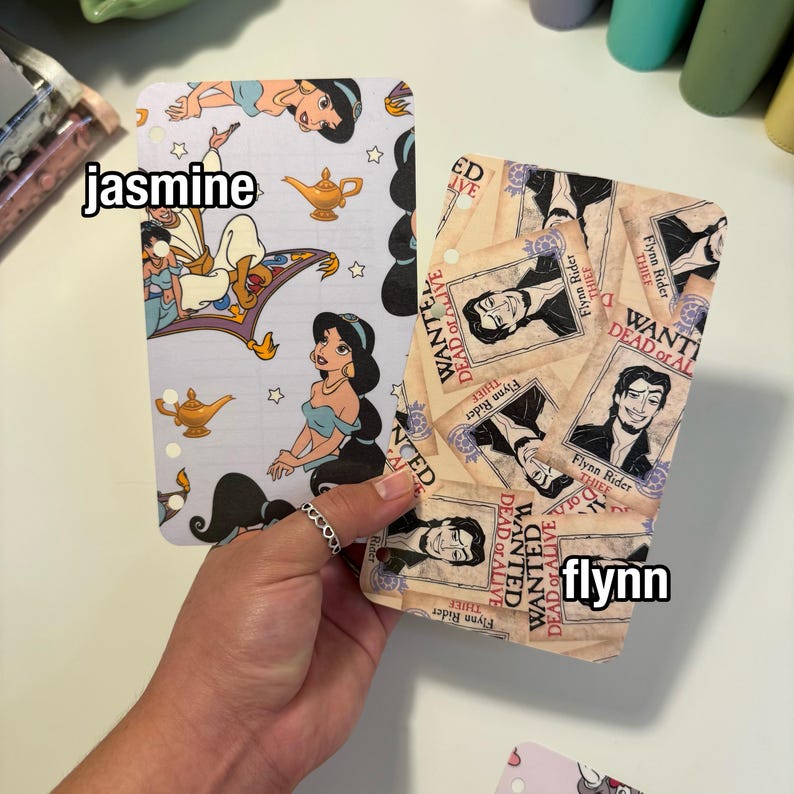 Op de afbeelding: Twee decoratieve plannerinzetstukken worden in een hand gehouden. E&eacute;n toont het personage Jasmine uit Aladdin, met de tekst "jasmine". De andere heeft een "Wanted Dead or Alive"-posterontwerp met de tekst "flynn".