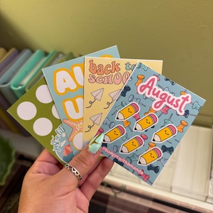 May include: Four colorful stickers with different designs. The first sticker is green with white dots. The second sticker is blue with a yellow star, a yellow sun, and the text "Just a little" in white. The third sticker is yellow with the text "Back to School" in pink and blue. The fourth sticker is blue with a pattern of pink pencils and white pushpins and the text "August" in pink.