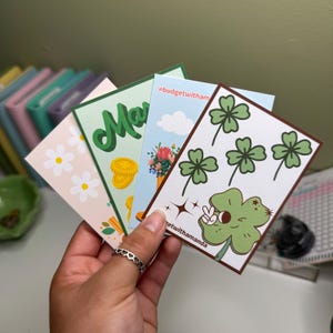 May include: Four different cards with various designs. One card features a green four-leaf clover with a cartoon character holding a peace sign. Another card has a green shamrock with the text "@budgetwithamanda". A third card has a green shamrock with the text "@budgetwitham". The fourth card has the word "March" in green letters with a gold coin design.