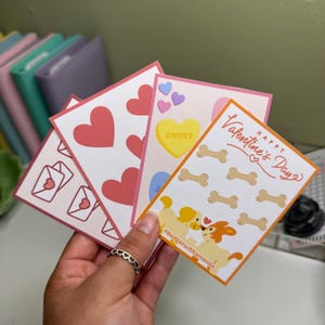 May include: Four Valentine's Day cards with different designs. One card has pink hearts, another has a candy heart with the word "SWEET", another has a dog couple with bones, and the last one has envelopes with hearts. The card with the dog couple has the text "Happy Valentine's Day" and the hashtag "#budgetwithamanda".
