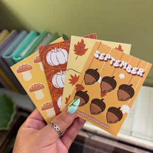 May include: Four autumn-themed cards with different designs. One card features a pumpkin and leaves, another has mushrooms, a third has acorns, and the last one says "SEPTEMBER" with acorns.
