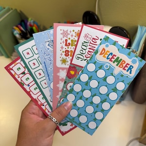 May include: A hand holds a fan of colorful budget tracker cards. The cards feature holiday themes with text like "December" and "Dear Santa." Each card has a different design, including snowflakes and Christmas trees. The cards are designed for financial planning.