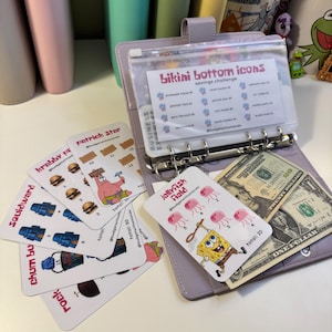 May include: A lilac binder with a clear pouch containing a savings challenge card titled "Bikini Bottom Icons." Several themed cards with cartoon characters and illustrations are scattered around the binder, along with US currency.
