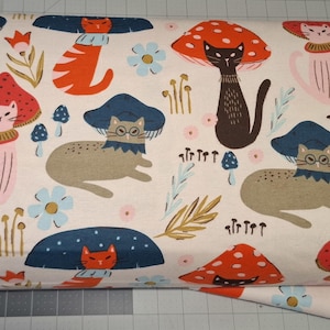 May include: Cream-coloured fabric featuring a playful design of cats, toadstools, and flowers. The cats are in different poses, some wearing hats. The toadstools are red and brown, and the flowers are blue and pink.