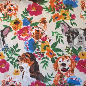 May include: A cream-colored fabric featuring a repeating pattern of various dog breeds and colorful flowers. The dogs include a French Bulldog, a Beagle, a Golden Retriever, and a Yorkshire Terrier, surrounded by pink, blue, orange, and yellow flowers.