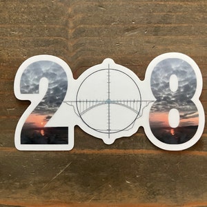 May include: A white sticker with the number 208 printed in black. The number 0 is replaced with a target graphic with a bridge in the center.